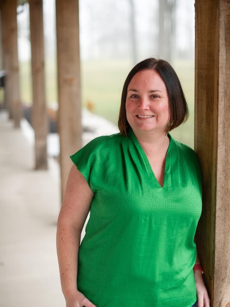 Portrait of Kari Cameron smiling, wearing a green blouse and leaning against a wooden pillar